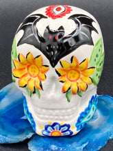 Load image into Gallery viewer, Inspired by nature, this ceramic skull statuette is full of vibrant color and intricate details. The primary color is white with a batty for bats theme and with multi-color floral motifs, along with hearts and leaves and lively colorful accents around the entire skull. Measurements: H 2 3/4” x W 2” x D 2”