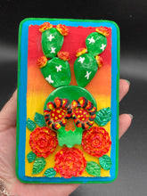 Load image into Gallery viewer, This handcrafted polymer clay wall art features a wood frame painted in a gradient from light yellow to dark red, creating a stunning sunset look. With a bright green skull cactus plant and bright yellow blooming flowers. Boasting vivid marigold blooms and a bright teal and green hue frame to make a bold contrast with the warm background hue. this piece is sure to make a statement. Measurements: