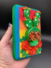Load image into Gallery viewer, This handcrafted polymer clay wall art features a wood frame painted in a gradient from light yellow to dark red, creating a stunning sunset look. With a bright green skull cactus plant and bright yellow blooming flowers. Boasting vivid marigold blooms and a bright teal and green hue frame to make a bold contrast with the warm background hue. this piece is sure to make a statement. Measurements: