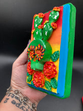 Load image into Gallery viewer, This handcrafted polymer clay wall art features a wood frame painted in a gradient from light yellow to dark red, creating a stunning sunset look. With a bright green skull cactus plant and bright yellow blooming flowers. Boasting vivid marigold blooms and a bright teal and green hue frame to make a bold contrast with the warm background hue. this piece is sure to make a statement. Measurements: