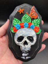 Load image into Gallery viewer, Showcasing a vibrant Frida Kahlo inspired calavera with a dark wood frame, this Day of the Dead wall decor piece is perfect for adding a traditional Mexican folk art touch to any home. Featuring an intricately detailed clay calavera in the style of Frida Kahlo, this Day of the Dead wall art decor is sure to be the centerpiece of any home décor. The calavera is wearing Frida's iconic crown of flowers and a monarch butterfly, ensuring it will bring both a unique and inspiring look to any wall.