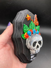 Load image into Gallery viewer, Showcasing a vibrant Frida Kahlo inspired calavera with a dark wood frame, this Day of the Dead wall decor piece is perfect for adding a traditional Mexican folk art touch to any home. Featuring an intricately detailed clay calavera in the style of Frida Kahlo, this Day of the Dead wall art decor is sure to be the centerpiece of any home décor. The calavera is wearing Frida's iconic crown of flowers and a monarch butterfly, ensuring it will bring both a unique and inspiring look to any wall.