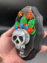 Load image into Gallery viewer, Showcasing a vibrant Frida Kahlo inspired calavera with a dark wood frame, this Day of the Dead wall decor piece is perfect for adding a traditional Mexican folk art touch to any home. Featuring an intricately detailed clay calavera in the style of Frida Kahlo, this Day of the Dead wall art decor is sure to be the centerpiece of any home décor. The calavera is wearing Frida's iconic crown of flowers and a monarch butterfly, ensuring it will bring both a unique and inspiring look to any wall.