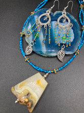 Load image into Gallery viewer, These large chandelier-style earrings feature blues, greens, and iridescent glass beads as well as a finely crafted silver plated leaf charm for added detail. Perfect for bohemian-inspired looks, these versatile earrings can elevate any wardrobe from whimsical hippie to boho chic. Measurements: 3 1/4” in length