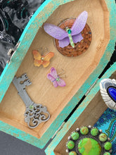 Load image into Gallery viewer, This Whimsical Fairy Wood Coffin Box is a truly unique and magical piece of home decor. With its vibrant turquoise exterior and eye-catching mini door knob, this Steampunk-inspired box is sure to be a conversation starter. Inside, the glittery gold interior and colorful butterfly detailing will bring a touch of fantasy to any room. The silver key and accompanying iridescent crystals complete the mystical and pastel goth look!