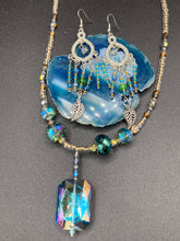 Load image into Gallery viewer, These large chandelier-style earrings feature blues, greens, and iridescent glass beads as well as a finely crafted silver plated leaf charm for added detail. Perfect for bohemian-inspired looks, these versatile earrings can elevate any wardrobe from whimsical hippie to boho chic. Measurements: 3 1/4” in length
