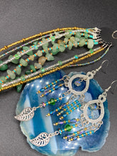 Load image into Gallery viewer, These large chandelier-style earrings feature blues, greens, and iridescent glass beads as well as a finely crafted silver plated leaf charm for added detail. Perfect for bohemian-inspired looks, these versatile earrings can elevate any wardrobe from whimsical hippie to boho chic. Measurements: 3 1/4” in length