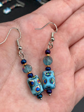 Load image into Gallery viewer, This set of glass beaded earrings features a vibrant blue shade with hints of purples and gold for an overall dynamic look. Its unique texture is derived from the combination of different size and shaped beads, resulting in a bold, eye-catching piece. For any discerning connoisseur, this set is a must-have addition to any jewelry collection. Measurements: 1 1/2” in length
