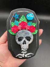 Load image into Gallery viewer, Showcasing a dark theme Frida Kahlo inspired calavera with a dark wood frame, this Day of the Dead wall decor piece is perfect for adding a traditional Mexican folk art touch to any home. Featuring an intricately detailed clay calavera in the style of Frida Kahlo, this Day of the Dead wall art decor is sure to be the centerpiece of any home décor. The calavera is wearing Frida's iconic crown of flowers, ensuring it will bring both a unique and inspiring look to any wall.