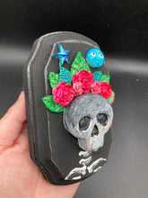 Load image into Gallery viewer, Showcasing a dark theme Frida Kahlo inspired calavera with a dark wood frame, this Day of the Dead wall decor piece is perfect for adding a traditional Mexican folk art touch to any home. Featuring an intricately detailed clay calavera in the style of Frida Kahlo, this Day of the Dead wall art decor is sure to be the centerpiece of any home décor. The calavera is wearing Frida's iconic crown of flowers, ensuring it will bring both a unique and inspiring look to any wall.
