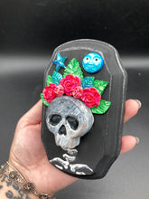 Load image into Gallery viewer, Showcasing a dark theme Frida Kahlo inspired calavera with a dark wood frame, this Day of the Dead wall decor piece is perfect for adding a traditional Mexican folk art touch to any home. Featuring an intricately detailed clay calavera in the style of Frida Kahlo, this Day of the Dead wall art decor is sure to be the centerpiece of any home décor. The calavera is wearing Frida's iconic crown of flowers, ensuring it will bring both a unique and inspiring look to any wall.