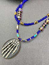 Load image into Gallery viewer, This spooky resin anatomical skeleton hand pendant is a perfect handy jewelry piece! The anatomical skeleton hand is made of resin with brass metal backing. Rich royal blue glass beads to give it a huge pop of color. Rectangular and hexagon rainbow hematite beads to compliment the shapes of the finger bones in the pendant. Bright canary yellow beads along the strands to break up the cool hues a bit. Necklace measures 16” adjustable up to 18” in length, pendant is 1 1/4”