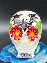 Load image into Gallery viewer, Inspired by nature, this ceramic skull statuette is full of vibrant color and intricate details. The primary color is white with a batty for bats theme and with multi-color floral motifs, along with hearts and leaves and lively colorful accents around the entire skull. Measurements: H 2 3/4” x W 2” x D 2”