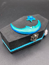 Load image into Gallery viewer, This sleek black box has an eye-catching turquoise crescent at its center, accompanied by a bright blue ribbon and a dainty white moon. Inside, the interior is decorated with a vivid royal blue and detailed with a small crescent moon and black-and-silver cross. Coffin Dimensions: Length: 4 1/2 Inches; Width: 2 1/2 inches; Height: 1 5/8 Inches