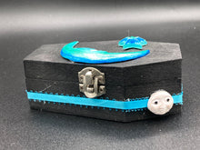 Load image into Gallery viewer, This sleek black box has an eye-catching turquoise crescent at its center, accompanied by a bright blue ribbon and a dainty white moon. Inside, the interior is decorated with a vivid royal blue and detailed with a small crescent moon and black-and-silver cross. Coffin Dimensions: Length: 4 1/2 Inches; Width: 2 1/2 inches; Height: 1 5/8 Inches