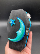 Load image into Gallery viewer, This sleek black box has an eye-catching turquoise crescent at its center, accompanied by a bright blue ribbon and a dainty white moon. Inside, the interior is decorated with a vivid royal blue and detailed with a small crescent moon and black-and-silver cross. Coffin Dimensions: Length: 4 1/2 Inches; Width: 2 1/2 inches; Height: 1 5/8 Inches