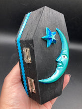 Load image into Gallery viewer, This sleek black box has an eye-catching turquoise crescent at its center, accompanied by a bright blue ribbon and a dainty white moon. Inside, the interior is decorated with a vivid royal blue and detailed with a small crescent moon and black-and-silver cross. Coffin Dimensions: Length: 4 1/2 Inches; Width: 2 1/2 inches; Height: 1 5/8 Inches