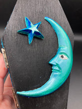 Load image into Gallery viewer, This sleek black box has an eye-catching turquoise crescent at its center, accompanied by a bright blue ribbon and a dainty white moon. Inside, the interior is decorated with a vivid royal blue and detailed with a small crescent moon and black-and-silver cross. Coffin Dimensions: Length: 4 1/2 Inches; Width: 2 1/2 inches; Height: 1 5/8 Inches