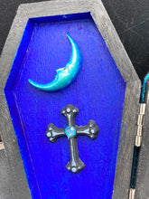Load image into Gallery viewer, This sleek black box has an eye-catching turquoise crescent at its center, accompanied by a bright blue ribbon and a dainty white moon. Inside, the interior is decorated with a vivid royal blue and detailed with a small crescent moon and black-and-silver cross. Coffin Dimensions: Length: 4 1/2 Inches; Width: 2 1/2 inches; Height: 1 5/8 Inches
