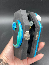Load image into Gallery viewer, This sleek black box has an eye-catching turquoise crescent at its center, accompanied by a bright blue ribbon and a dainty white moon. Inside, the interior is decorated with a vivid royal blue and detailed with a small crescent moon and black-and-silver cross. Coffin Dimensions: Length: 4 1/2 Inches; Width: 2 1/2 inches; Height: 1 5/8 Inches
