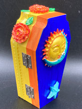 Load image into Gallery viewer, This coffin-shaped box features a bright royal blue exterior, with a bold sun and crescent adorning the center. Complemented with marigold flowers, the inside is painted in a bright red hue with turquoise and yellow cross detailing and traditional Mexican-themed fabric. Coffin Dimensions: Length: 4 1/2 Inches; Width: 2 1/2 inches; Height: 1 5/8 Inches