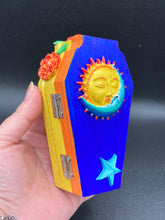 Load image into Gallery viewer, This coffin-shaped box features a bright royal blue exterior, with a bold sun and crescent adorning the center. Complemented with marigold flowers, the inside is painted in a bright red hue with turquoise and yellow cross detailing and traditional Mexican-themed fabric. Coffin Dimensions: Length: 4 1/2 Inches; Width: 2 1/2 inches; Height: 1 5/8 Inches