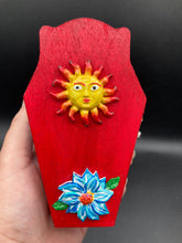 Load image into Gallery viewer, This coffin-shaped box features a bright red exterior, with a bold yellow, orange and red sun adorning the center. Complemented with a shimmery turquoise-blue ribbon and flower, the inside is painted in a dark royal blue hue with turquoise crescent moon detailing and traditional Mexican-themed fabric. Coffin Dimensions: Length: 4 1/2 Inches; Width: 2 1/2 inches; Height: 1 5/8 Inches