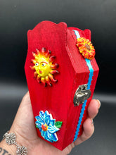 Load image into Gallery viewer, This coffin-shaped box features a bright red exterior, with a bold yellow, orange and red sun adorning the center. Complemented with a shimmery turquoise-blue ribbon and flower, the inside is painted in a dark royal blue hue with turquoise crescent moon detailing and traditional Mexican-themed fabric. Coffin Dimensions: Length: 4 1/2 Inches; Width: 2 1/2 inches; Height: 1 5/8 Inches