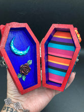 Load image into Gallery viewer, This coffin-shaped box features a bright red exterior, with a bold yellow, orange and red sun adorning the center. Complemented with a shimmery turquoise-blue ribbon and flower, the inside is painted in a dark royal blue hue with turquoise crescent moon detailing and traditional Mexican-themed fabric. Coffin Dimensions: Length: 4 1/2 Inches; Width: 2 1/2 inches; Height: 1 5/8 Inches