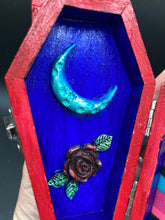 Load image into Gallery viewer, This coffin-shaped box features a bright red exterior, with a bold yellow, orange and red sun adorning the center. Complemented with a shimmery turquoise-blue ribbon and flower, the inside is painted in a dark royal blue hue with turquoise crescent moon detailing and traditional Mexican-themed fabric. Coffin Dimensions: Length: 4 1/2 Inches; Width: 2 1/2 inches; Height: 1 5/8 Inches
