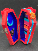 Load image into Gallery viewer, This coffin-shaped box features a bright red exterior, with a bold yellow, orange and red sun adorning the center. Complemented with a shimmery turquoise-blue ribbon and flower, the inside is painted in a dark royal blue hue with turquoise crescent moon detailing and traditional Mexican-themed fabric. Coffin Dimensions: Length: 4 1/2 Inches; Width: 2 1/2 inches; Height: 1 5/8 Inches