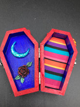 Load image into Gallery viewer, This coffin-shaped box features a bright red exterior, with a bold yellow, orange and red sun adorning the center. Complemented with a shimmery turquoise-blue ribbon and flower, the inside is painted in a dark royal blue hue with turquoise crescent moon detailing and traditional Mexican-themed fabric. Coffin Dimensions: Length: 4 1/2 Inches; Width: 2 1/2 inches; Height: 1 5/8 Inches