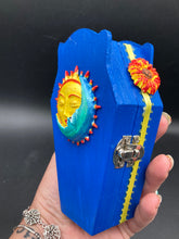 Load image into Gallery viewer, This coffin-shaped box features a bright royal blue exterior, with a bold sun and crescent adorning the center. Complemented with a shimmery pastel-yellow ribbon and flower, the inside is painted in a canary yellow hue with turquoise crescent moon detailing and traditional Mexican-themed fabric. Coffin Dimensions: Length: 4 1/2 Inches; Width: 2 1/2 inches; Height: 1 5/8 Inches