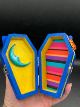 Load image into Gallery viewer, This coffin-shaped box features a bright royal blue exterior, with a bold sun and crescent adorning the center. Complemented with a shimmery pastel-yellow ribbon and flower, the inside is painted in a canary yellow hue with turquoise crescent moon detailing and traditional Mexican-themed fabric. Coffin Dimensions: Length: 4 1/2 Inches; Width: 2 1/2 inches; Height: 1 5/8 Inches