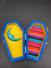 Load image into Gallery viewer, This coffin-shaped box features a bright royal blue exterior, with a bold sun and crescent adorning the center. Complemented with a shimmery pastel-yellow ribbon and flower, the inside is painted in a canary yellow hue with turquoise crescent moon detailing and traditional Mexican-themed fabric. Coffin Dimensions: Length: 4 1/2 Inches; Width: 2 1/2 inches; Height: 1 5/8 Inches