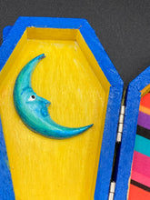 Load image into Gallery viewer, This coffin-shaped box features a bright royal blue exterior, with a bold sun and crescent adorning the center. Complemented with a shimmery pastel-yellow ribbon and flower, the inside is painted in a canary yellow hue with turquoise crescent moon detailing and traditional Mexican-themed fabric. Coffin Dimensions: Length: 4 1/2 Inches; Width: 2 1/2 inches; Height: 1 5/8 Inches