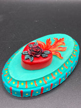 Load image into Gallery viewer, This Sacred Heart Wall Art Decor is the perfect way to enhance your home with an artistic flair. This beautiful artwork features a bright turquoise wooden frame with a hand-crafted bright red heart. The heart is detailed with dark red rose and vibrant green leaves, with a vibrant fire embellishment over the center. Gold decorative embellishments at the sides of the heart add a luxurious touch. H 5 3/8" x W 3 3/8” x D 1 1/2"