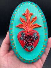 Load image into Gallery viewer, This Sacred Heart Wall Art Decor is the perfect way to enhance your home with an artistic flair. This beautiful artwork features a bright turquoise wooden frame with a hand-crafted bright red heart. The heart is detailed with dark red rose and vibrant green leaves, with a vibrant fire embellishment over the center. Gold decorative embellishments at the sides of the heart add a luxurious touch. H 5 3/8" x W 3 3/8” x D 1 1/2"