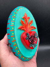 Load image into Gallery viewer, This Sacred Heart Wall Art Decor is the perfect way to enhance your home with an artistic flair. This beautiful artwork features a bright turquoise wooden frame with a hand-crafted bright red heart. The heart is detailed with dark red rose and vibrant green leaves, with a vibrant fire embellishment over the center. Gold decorative embellishments at the sides of the heart add a luxurious touch. H 5 3/8" x W 3 3/8” x D 1 1/2"
