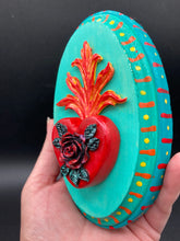 Load image into Gallery viewer, This Sacred Heart Wall Art Decor is the perfect way to enhance your home with an artistic flair. This beautiful artwork features a bright turquoise wooden frame with a hand-crafted bright red heart. The heart is detailed with dark red rose and vibrant green leaves, with a vibrant fire embellishment over the center. Gold decorative embellishments at the sides of the heart add a luxurious touch. H 5 3/8" x W 3 3/8” x D 1 1/2"