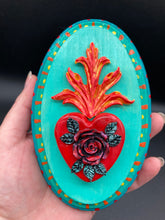 Load image into Gallery viewer, This Sacred Heart Wall Art Decor is the perfect way to enhance your home with an artistic flair. This beautiful artwork features a bright turquoise wooden frame with a hand-crafted bright red heart. The heart is detailed with dark red rose and vibrant green leaves, with a vibrant fire embellishment over the center. Gold decorative embellishments at the sides of the heart add a luxurious touch. H 5 3/8" x W 3 3/8” x D 1 1/2"