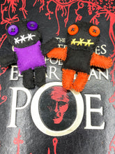 Load image into Gallery viewer, Short and tiny black and purple Voodoo Doll is 3" in height. Torso is purple while legs and arms and head are all black like the night. He has matching purple buttons for eyes and has a sowed shut mouth. Short and tiny black and orange Voodoo Doll is 3" in height. Torso and head is all black while legs and arms are bright orange, perfect Halloween colors. He has matching bright orange buttons for eyes and has a sowed shut mouth.