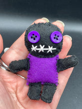 Load image into Gallery viewer, Short and tiny black and purple Voodoo Doll is 3" in height. Torso is purple while legs and arms and head are all black like the night. He has matching purple buttons for eyes and has a sowed shut mouth.