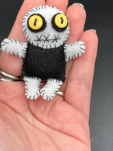 Load image into Gallery viewer, Super tiny black and white Voodoo Doll has a black torso and white arms, legs and big round white head. He has bulging yellow button eyes, too big for his tiny head. He looks like he’s almost smiling…almost. He stand at 2 1/2" in height