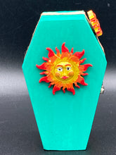 Load image into Gallery viewer, This coffin-shaped box features a bright turquoise exterior, with a bold yellow, orange and red sun adorning the center. Complemented with a shimmery rose-gold ribbon and flower, the inside is painted in a vibrant orange hue with turquoise crescent moon detailing and traditional Mexican-themed fabric. Coffin Dimensions: Length: 4 1/2 Inches; Width: 2 1/2 inches; Height: 1 5/8 Inches