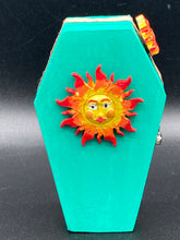 Load image into Gallery viewer, This coffin-shaped box features a bright turquoise exterior, with a bold yellow, orange and red sun adorning the center. Complemented with a shimmery rose-gold ribbon and flower, the inside is painted in a vibrant orange hue with turquoise crescent moon detailing and traditional Mexican-themed fabric. Coffin Dimensions: Length: 4 1/2 Inches; Width: 2 1/2 inches; Height: 1 5/8 Inches
