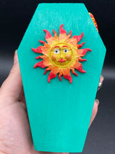 Load image into Gallery viewer, This coffin-shaped box features a bright turquoise exterior, with a bold yellow, orange and red sun adorning the center. Complemented with a shimmery rose-gold ribbon and flower, the inside is painted in a vibrant orange hue with turquoise crescent moon detailing and traditional Mexican-themed fabric. Coffin Dimensions: Length: 4 1/2 Inches; Width: 2 1/2 inches; Height: 1 5/8 Inches
