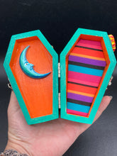 Load image into Gallery viewer, This coffin-shaped box features a bright turquoise exterior, with a bold yellow, orange and red sun adorning the center. Complemented with a shimmery rose-gold ribbon and flower, the inside is painted in a vibrant orange hue with turquoise crescent moon detailing and traditional Mexican-themed fabric. Coffin Dimensions: Length: 4 1/2 Inches; Width: 2 1/2 inches; Height: 1 5/8 Inches
