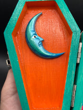 Load image into Gallery viewer, This coffin-shaped box features a bright turquoise exterior, with a bold yellow, orange and red sun adorning the center. Complemented with a shimmery rose-gold ribbon and flower, the inside is painted in a vibrant orange hue with turquoise crescent moon detailing and traditional Mexican-themed fabric. Coffin Dimensions: Length: 4 1/2 Inches; Width: 2 1/2 inches; Height: 1 5/8 Inches