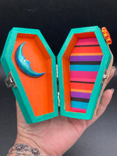 Load image into Gallery viewer, This coffin-shaped box features a bright turquoise exterior, with a bold yellow, orange and red sun adorning the center. Complemented with a shimmery rose-gold ribbon and flower, the inside is painted in a vibrant orange hue with turquoise crescent moon detailing and traditional Mexican-themed fabric. Coffin Dimensions: Length: 4 1/2 Inches; Width: 2 1/2 inches; Height: 1 5/8 Inches